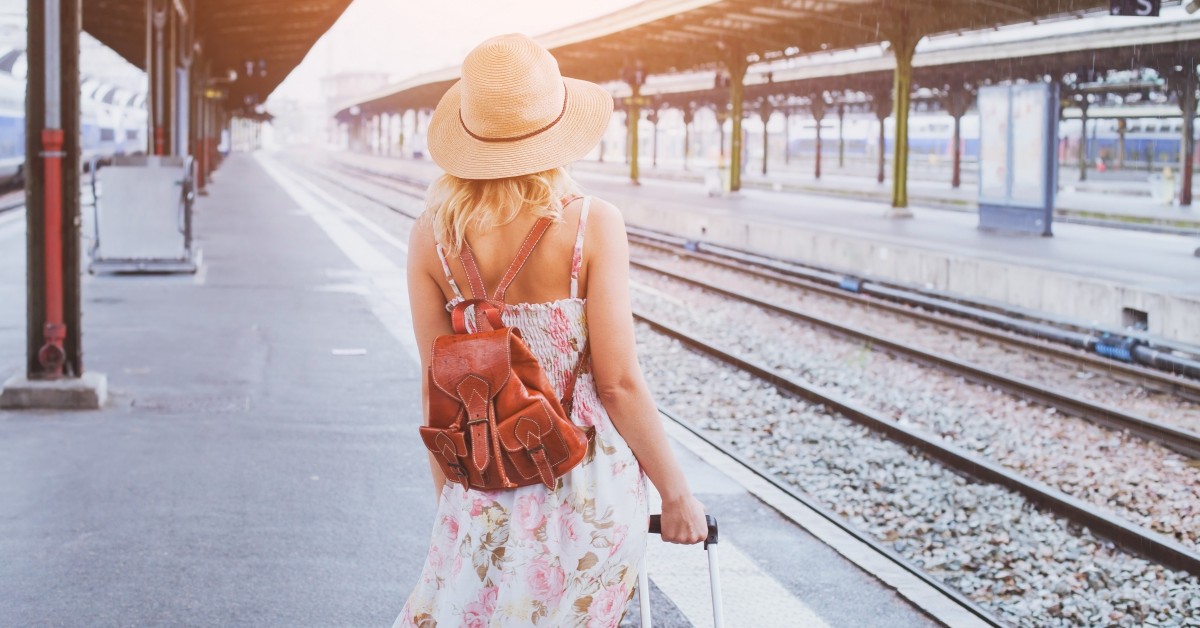 Blonde woman with red backpack and rolling luggage waiting for train