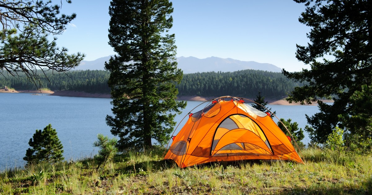 Orange camping tent on hill overlooking trees, mountain, and lake