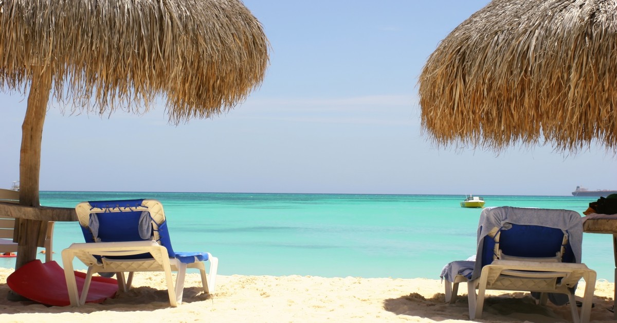 Tranquil scene of two beach chairs under palm trees overlooking clear blue ocean waters with boat in background