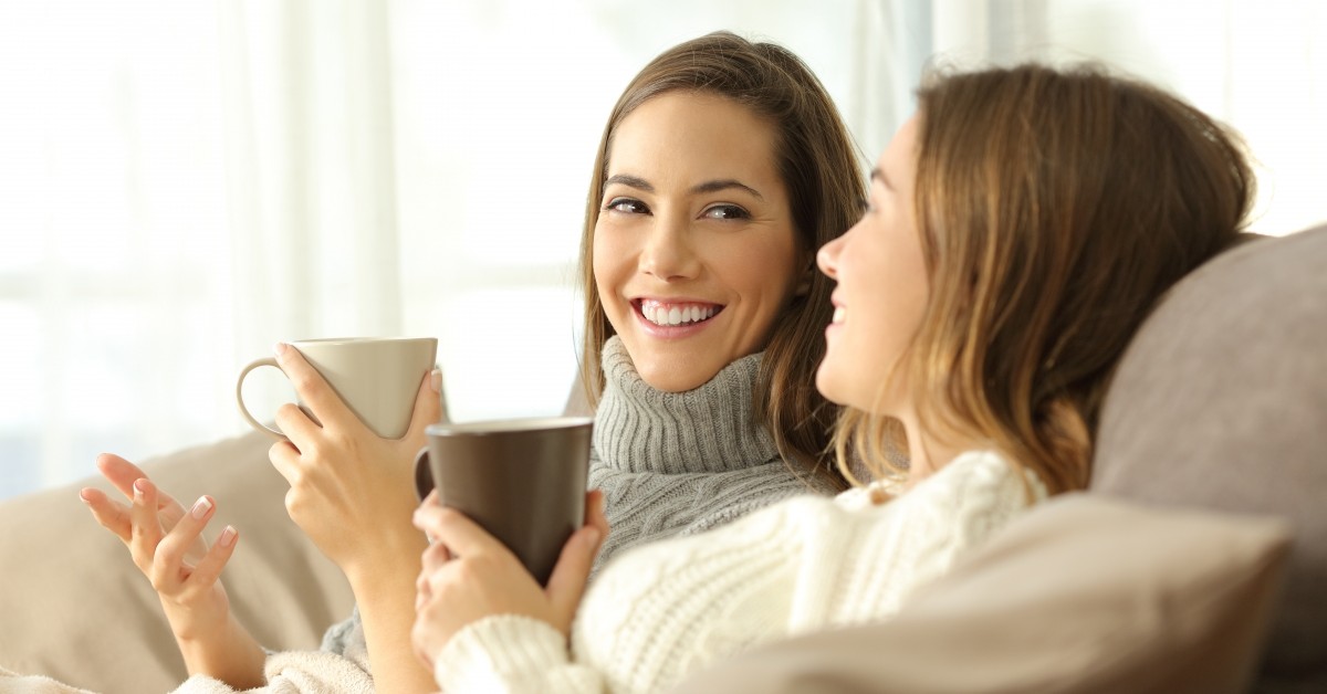 Two smiling young women sitting on a couch talking and drinking coffee