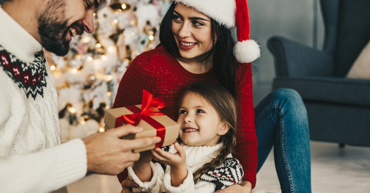 Mother and father giving their young daughter a wrapped Christmas gift with home holiday decor in background.