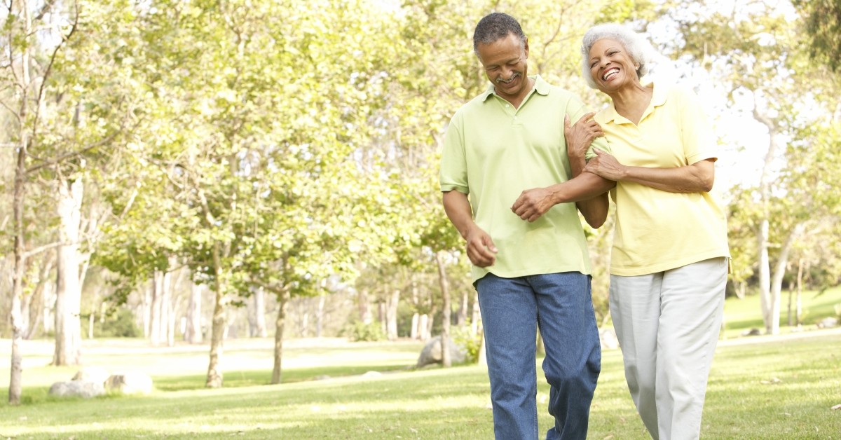 Elderly couple walking outside smiling arm-in-arm with trees in background.