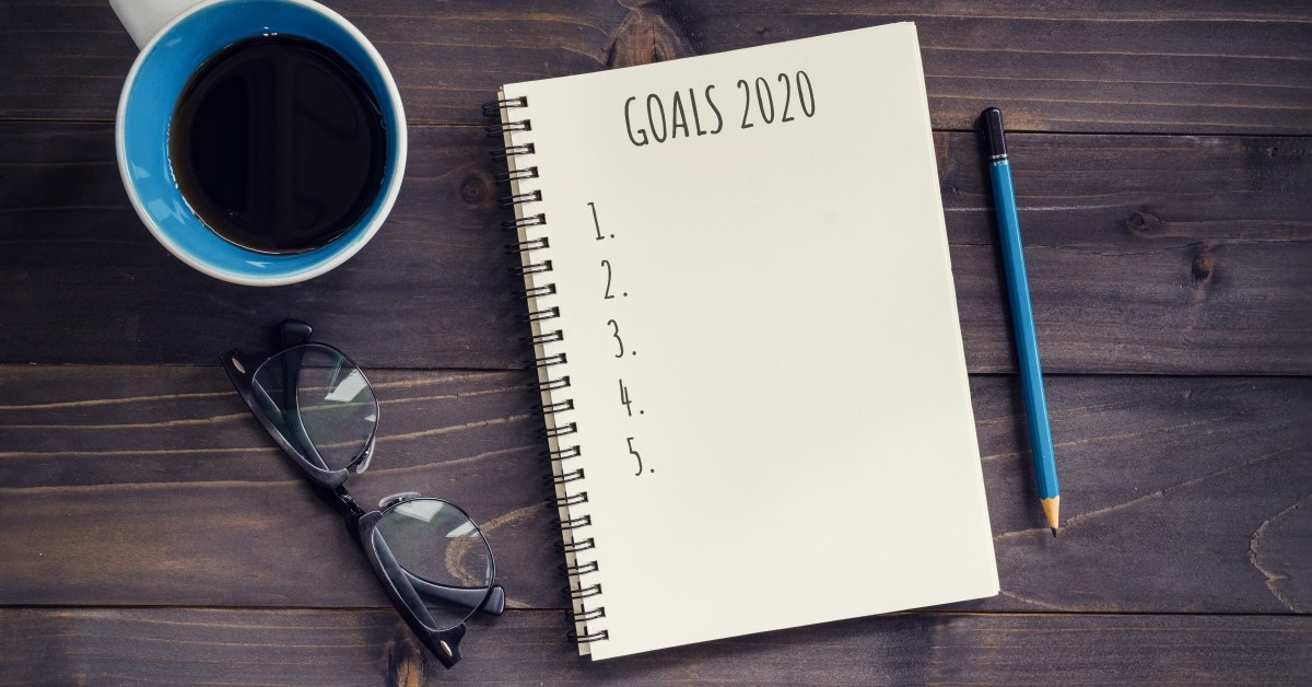 Aerial, close-up view of notepad, coffee cup, pen and eye-glasses with notebook text, "Goals: 1, 2, 3, 4, 5"