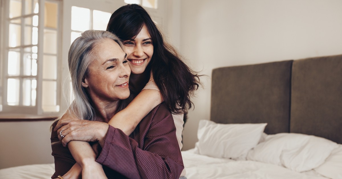 Middle-aged mother with teenage daughter embracing from behind, sitting in bedroom smiling