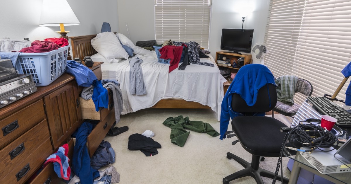 Scene of messy bedroom with laundry strewn about and desk with keyboard.