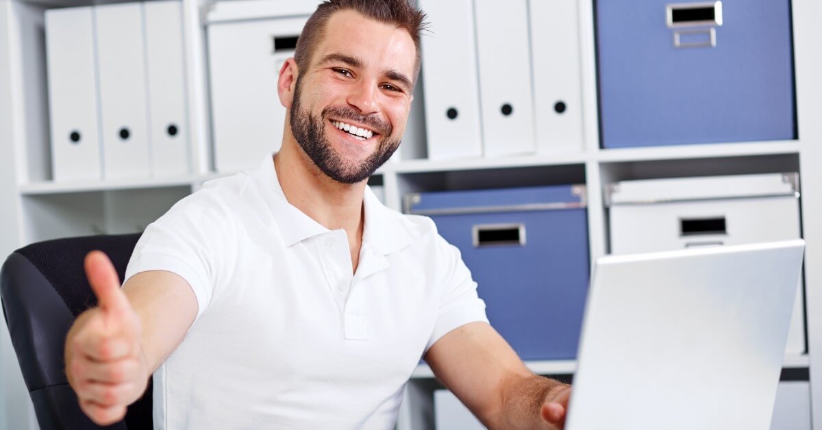 Man at desk giving thumbs up to camera in office setting.