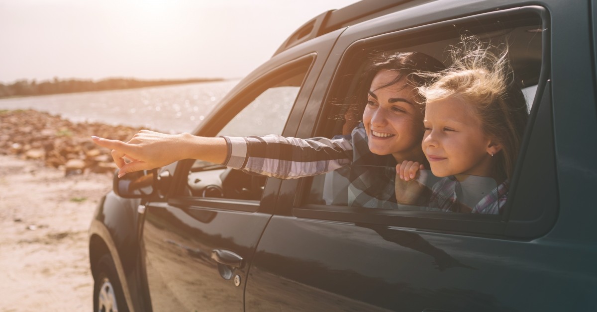 Woman and young girl looking out of the back passenger window of a car and smiling, with beach in background