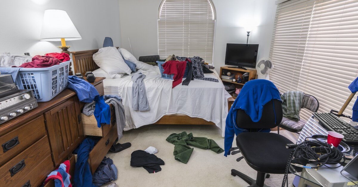 Scene of messy bedroom with laundry strewn about and desk with keyboard.