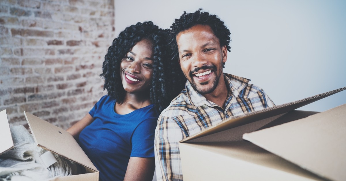 Smiling man and woman holding moving boxes in room with exposed brick wall